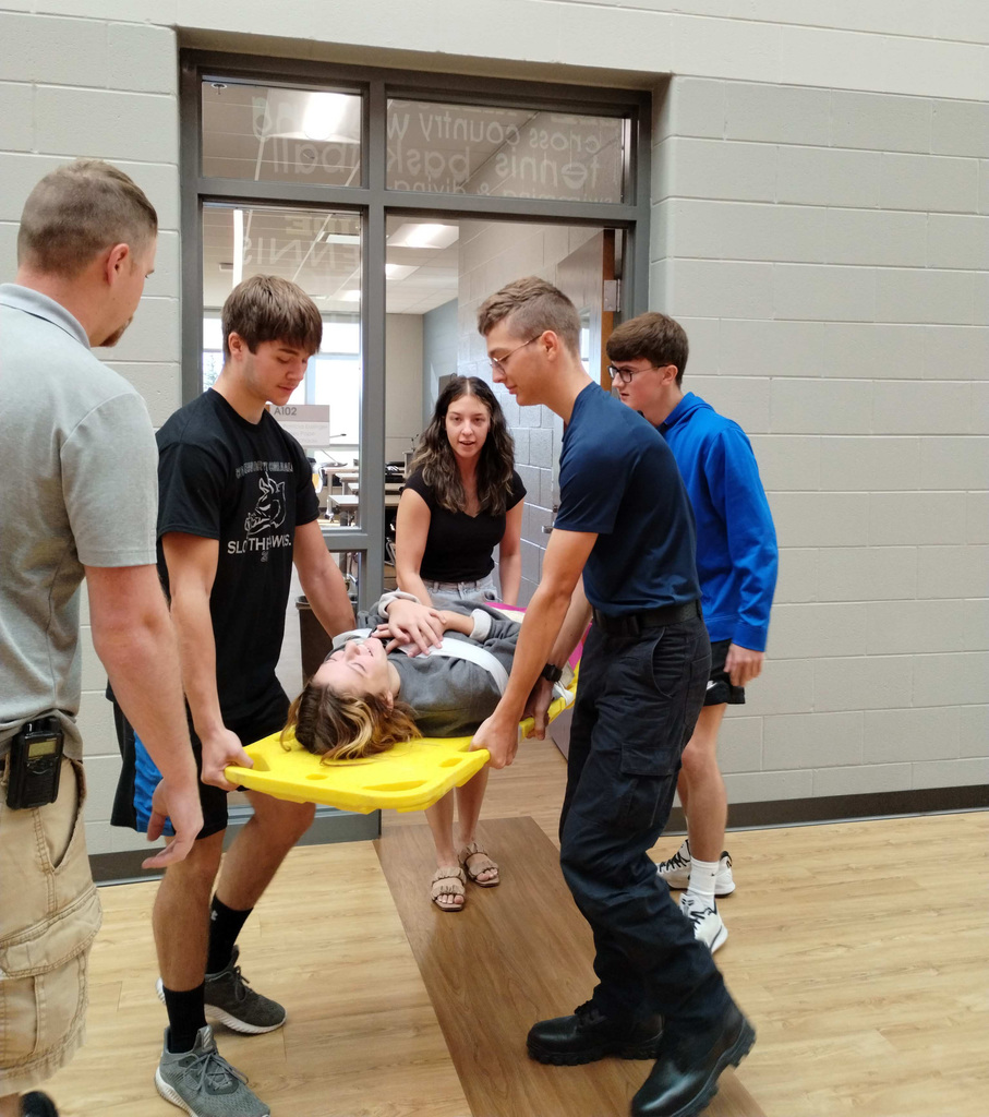 Five students hold onto a yellow stretcher with another student lying on the stretcher for practice safe lifting.