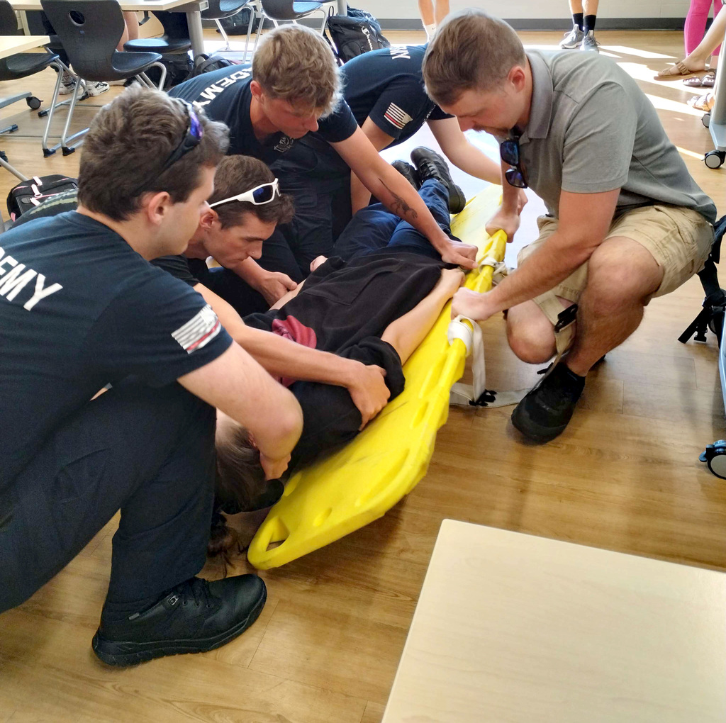 Student kneel over a classmate as they practice safely moving a patient onto a yellow stretcher. 