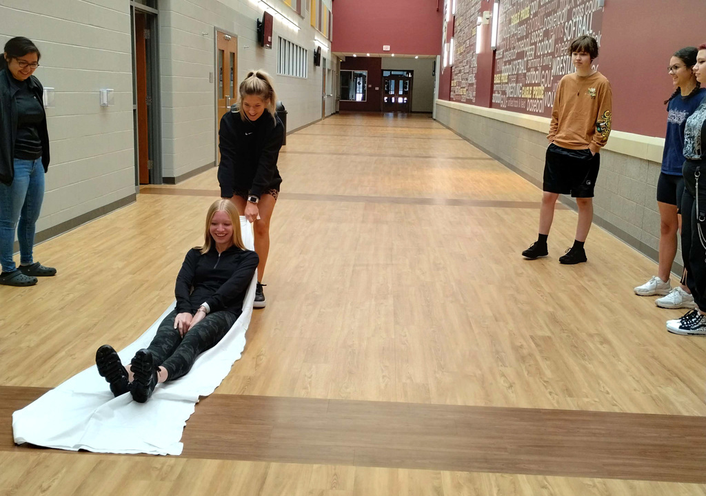 A student pulls a sheet with another student on it down the hall while students observe around them.