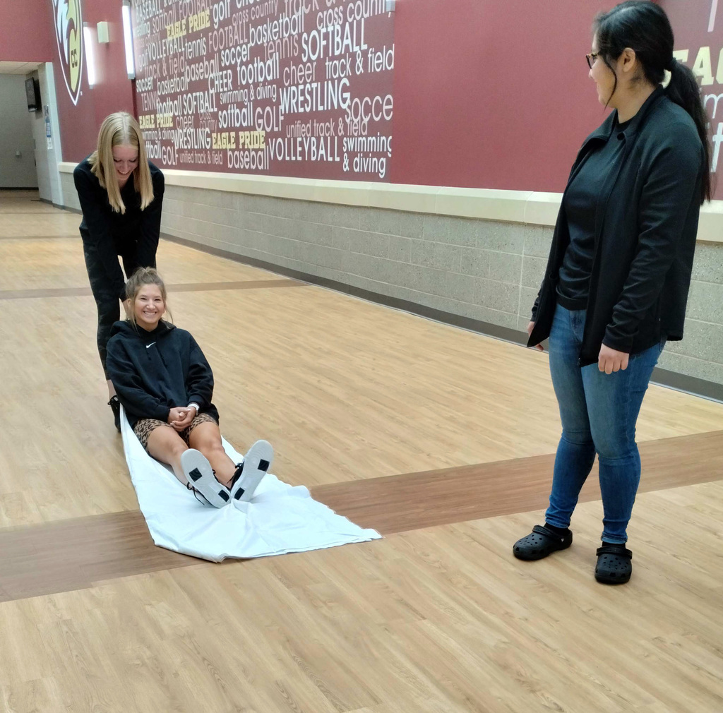 A student practices safely pulling and moving another student on a sheet while another student watches from the right side of the screen. 